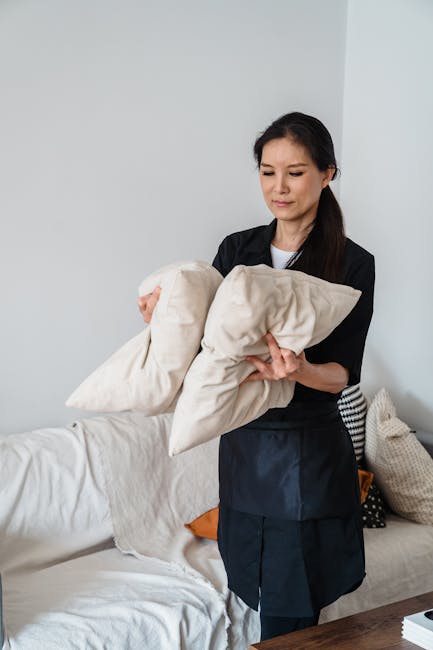 A woman in black attire holds two beige cushions while standing in a living room with a white sofa that has a light-colored cover, seen near a stack of cushions in different textures and colors. The r